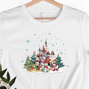 May include: White t-shirt with a Christmas-themed graphic featuring Mickey Mouse and friends in front of a snow-covered castle. The graphic includes a Christmas tree, presents, and snowflakes.