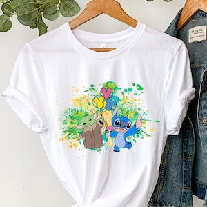 K&ouml;nnte beinhalten: Wei&szlig;es T-Shirt mit einer bunten Grafik von Stitch und Baby Yoda, die Ballons halten. Das Shirt h&auml;ngt an einem Kleiderb&uuml;gel mit einer Jeansjacke und einem Strohhut.