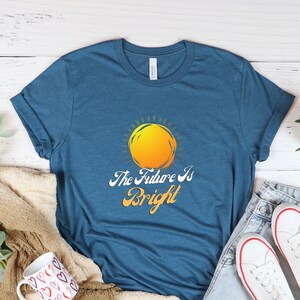 May include: A teal blue t-shirt with a yellow sun graphic and the text "The Future Is Bright". The shirt is laid out on a white wooden surface with a brown blanket, a white mug with red hearts, a potted plant, and a pair of white sneakers.