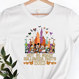 May include: White t-shirt featuring a colourful graphic of Mickey Mouse and his friends in front of a castle. The text "Not So Scary Halloween Party! 2023" is printed on the shirt.