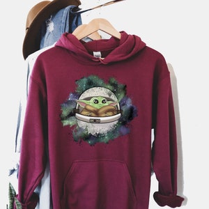 May include: Burgundy hooded sweatshirt with a graphic print of a green alien character in a spaceship against a galaxy background.