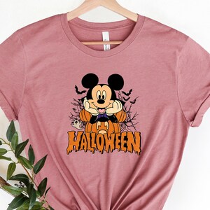 K&ouml;nnte beinhalten: Ein mauvefarbenes T-Shirt mit einem Aufdruck von Mickey Mouse, der als Vampir verkleidet ist und einen K&uuml;rbis h&auml;lt. Der Text "HALLOWEEN" ist unter Mickey Mouse aufgedruckt.