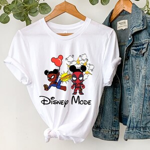 May include: White t-shirt with a graphic of Mickey Mouse dressed as Spider-Man and Deadpool. The graphic includes the words "POW!" and "Disney Mode".