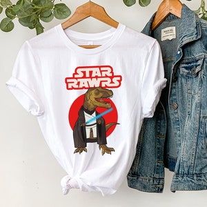 May include: White t-shirt with the text "STAR RAWRS" in red and a cartoon dinosaur with a lightsaber. A denim jacket and a straw hat hang nearby. The shirt is on a wooden hanger.