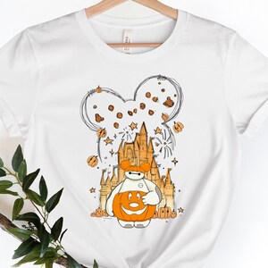 May include: White t-shirt with a cartoon illustration of a robot wearing a pumpkin for a head. The robot is standing in front of a castle with fireworks and Mickey Mouse ears in the background.