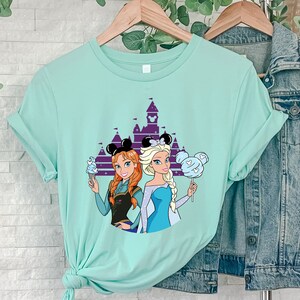 以下が含まれることがあります： ディズニーの城、アナとエルサ（アナと雪の女王）のグラフィックと「ディズニー」の文字がプリントされたターコイズブルーのTシャツ。アナはアイスクリームコーンを持っており、エルサは綿菓子を持っています。