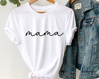 May include: White t-shirt with the word "mama" written in black cursive script. The shirt is hanging on a hanger with a denim jacket and a straw hat.