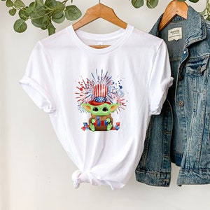 May include: White t-shirt with a green character wearing a red, white, and blue hat and holding a red, white, and blue flag. The character is surrounded by red, white, and blue fireworks. The t-shirt is on a hanger with a denim jacket next to it.