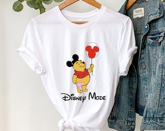Könnte beinhalten: Weißes T-Shirt mit einem Winnie-the-Pooh-Motiv, das einen roten Ballon mit Mickey-Maus-Ohren hält. Der Text "Disney Mode" ist unter dem Motiv gedruckt.