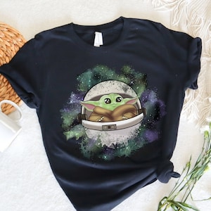 May include: Black t-shirt with a green alien character in a space suit against a galaxy background.