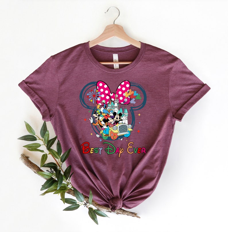 May include: A maroon t-shirt with a graphic of Mickey Mouse and friends in a car in front of Cinderella's Castle. The graphic is inside a Mickey Mouse ear silhouette with a pink polka dot bow. The text "Best Day Ever" is below the graphic.