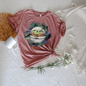 May include: A mauve t-shirt with a graphic print of a green alien character in a white space helmet. The alien is surrounded by a galaxy-like design with green, blue, and purple colors.