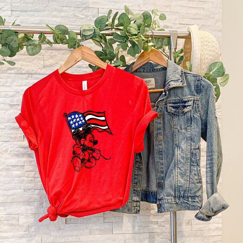 Puede incluir: Camiseta roja con un gr&aacute;fico de Mickey Mouse sosteniendo una bandera estadounidense. La camiseta est&aacute; en una percha con una chaqueta vaquera.