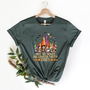 May include: A green t-shirt featuring a graphic of Mickey Mouse and his friends in front of a castle. The text "Not So Scary Halloween Party! 2023" is printed on the shirt.
