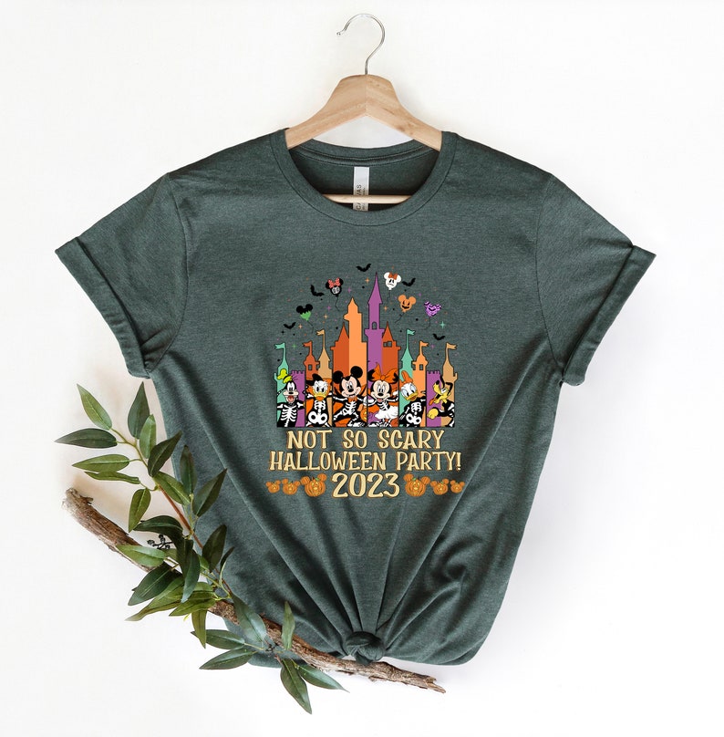 May include: A green t-shirt featuring a graphic of Mickey Mouse and his friends in front of a castle. The text "Not So Scary Halloween Party! 2023" is printed on the shirt.