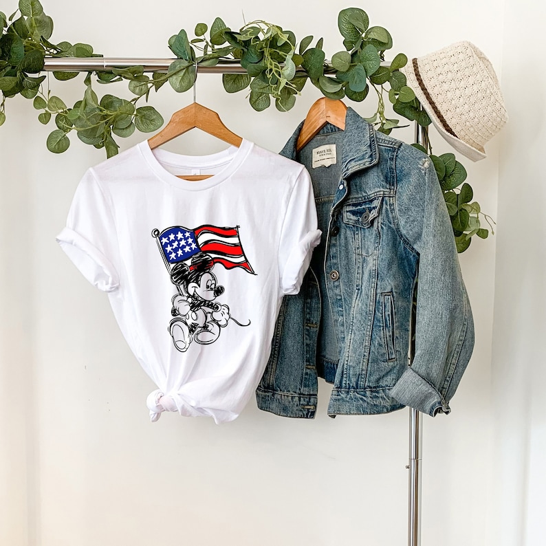 Puede incluir: Camiseta blanca con una ilustraci&oacute;n en blanco y negro de Mickey Mouse sosteniendo una bandera estadounidense. La bandera es roja, blanca y azul.