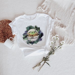 May include: White t-shirt with a green alien character in a spaceship, surrounded by a watercolor galaxy design.