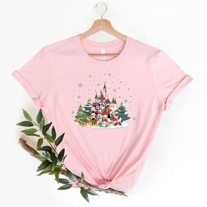 May include: Pink t-shirt with a Christmas-themed design featuring Mickey Mouse and friends in front of a castle with snow falling.