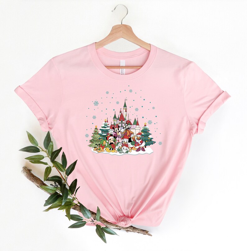 May include: Pink t-shirt with a Christmas-themed design featuring Mickey Mouse and friends in front of a castle with snow falling.