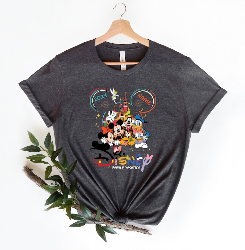 Puede incluir: Una camiseta gris oscuro con un gr&aacute;fico colorido de Mickey Mouse, Minnie Mouse, Donald Duck, Goofy y Pluto frente al Castillo de Cenicienta. El gr&aacute;fico tambi&eacute;n incluye el texto "Disney Family Vacation" y el a&ntilde;o "2024".