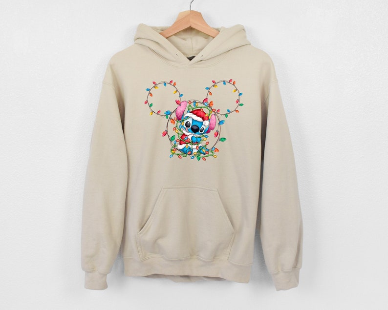 Może przedstawiać: Beżowa bluza z kapturem z niebieską postacią Stitch, kt&oacute;ra ma czapkę Świętego Mikołaja i jest opleciona świątecznymi lampkami. Lampki są ułożone w kształcie uszu Myszki Miki.