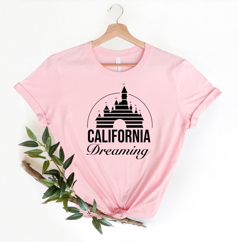 Puede incluir: Camiseta rosa con una silueta negra del castillo de la Bella Durmiente y el texto "CALIFORNIA Dreaming" impreso en la parte delantera.