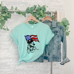 Puede incluir: Una camiseta azul claro con una imagen de dibujos animados en blanco y negro de Mickey Mouse sosteniendo una bandera estadounidense. La bandera es roja, blanca y azul.