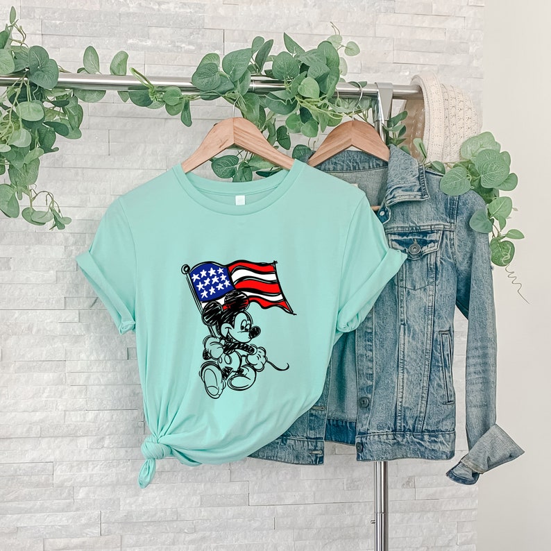 Puede incluir: Una camiseta azul claro con una imagen de dibujos animados en blanco y negro de Mickey Mouse sosteniendo una bandera estadounidense. La bandera es roja, blanca y azul.