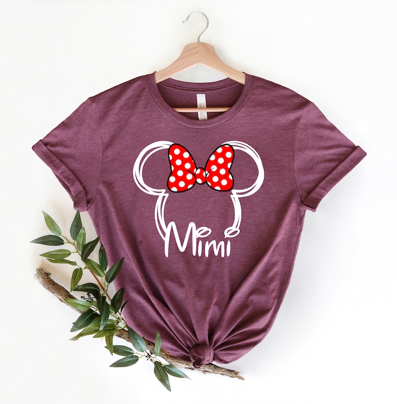 Op de afbeelding: Een maroon t-shirt met een witte omtrek van Mickey Mouse oren en een rode strik met witte stippen. De tekst "Mimi" staat onder de oren.