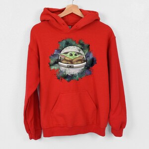 May include: Red hooded sweatshirt with a graphic print of a green alien character in a spaceship against a galaxy background.