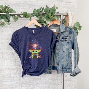 May include: A navy blue t-shirt with a graphic of a green character wearing a red, white, and blue hat with fireworks in the background. The shirt is hanging on a hanger with a denim jacket.