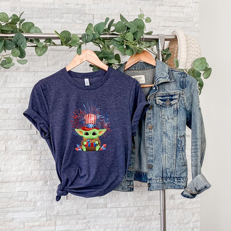 May include: A navy blue t-shirt with a graphic of a green character wearing a red, white, and blue hat with fireworks in the background. The shirt is hanging on a hanger with a denim jacket.