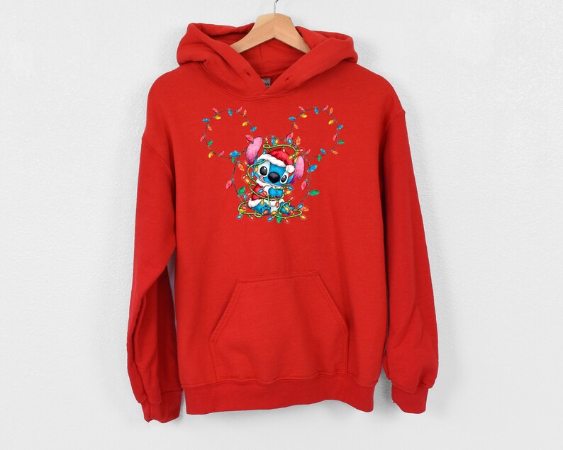 Może przedstawiać: Czerwona bluza z kapturem z niebieskim bohaterem Stitch, kt&oacute;ry nosi czapkę Świętego Mikołaja i jest zaplątany w świąteczne lampki.