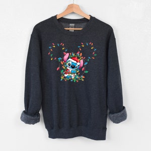 Może przedstawiać: Szara bluza z ciemnym odcieniem z niebieską postacią z kresk&oacute;wek zaplątaną w kolorowe świąteczne lampki. Postać ma na sobie czapkę Świętego Mikołaja i trzyma sznur lampek.