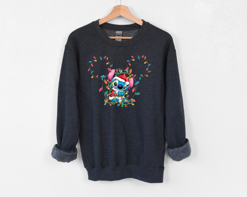 Może przedstawiać: Szara bluza z ciemnym odcieniem z niebieską postacią z kresk&oacute;wek zaplątaną w kolorowe świąteczne lampki. Postać ma na sobie czapkę Świętego Mikołaja i trzyma sznur lampek.