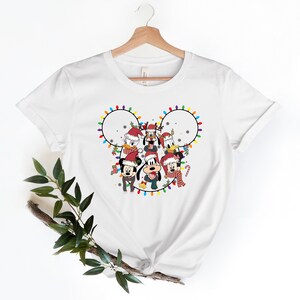 May include: White t-shirt with a Christmas-themed design featuring Mickey Mouse and his friends. The design shows Mickey, Minnie, Donald, Goofy, Pluto, and Daisy in Santa hats, surrounded by colorful Christmas lights in the shape of Mickey Mouse's ears.