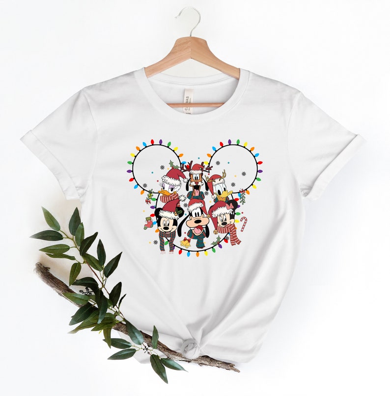 May include: White t-shirt with a Christmas-themed design featuring Mickey Mouse and his friends. The design shows Mickey, Minnie, Donald, Goofy, Pluto, and Daisy in Santa hats, surrounded by colorful Christmas lights in the shape of Mickey Mouse's ears.