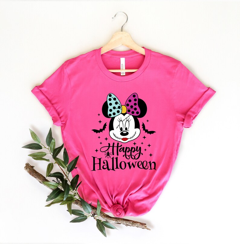 Puede incluir: Camiseta rosa con una ilustraci&oacute;n en blanco y negro de un rat&oacute;n de dibujos animados con una sonrisa cosida, que lleva un lazo azul y morado. El texto "Happy Halloween" est&aacute; impreso debajo del rat&oacute;n.