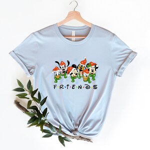 May include: Light blue t-shirt with a graphic of Mickey Mouse, Donald Duck, Goofy, and Pluto wearing Santa hats and green scarves. The text "FRIENDS" is below the characters.