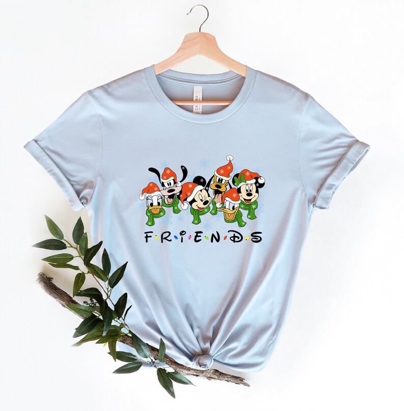 May include: Light blue t-shirt with a graphic of Mickey Mouse, Donald Duck, Goofy, and Pluto wearing Santa hats and green scarves. The text "FRIENDS" is below the characters.