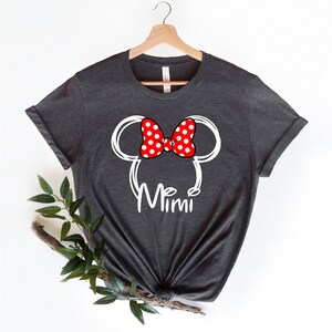 Op de afbeelding: Een donkergrijs T-shirt met een witte omtrek van Mickey Mouse oren en een rode strik met witte stippen. De tekst "Mimi" is onder de oren gedrukt.