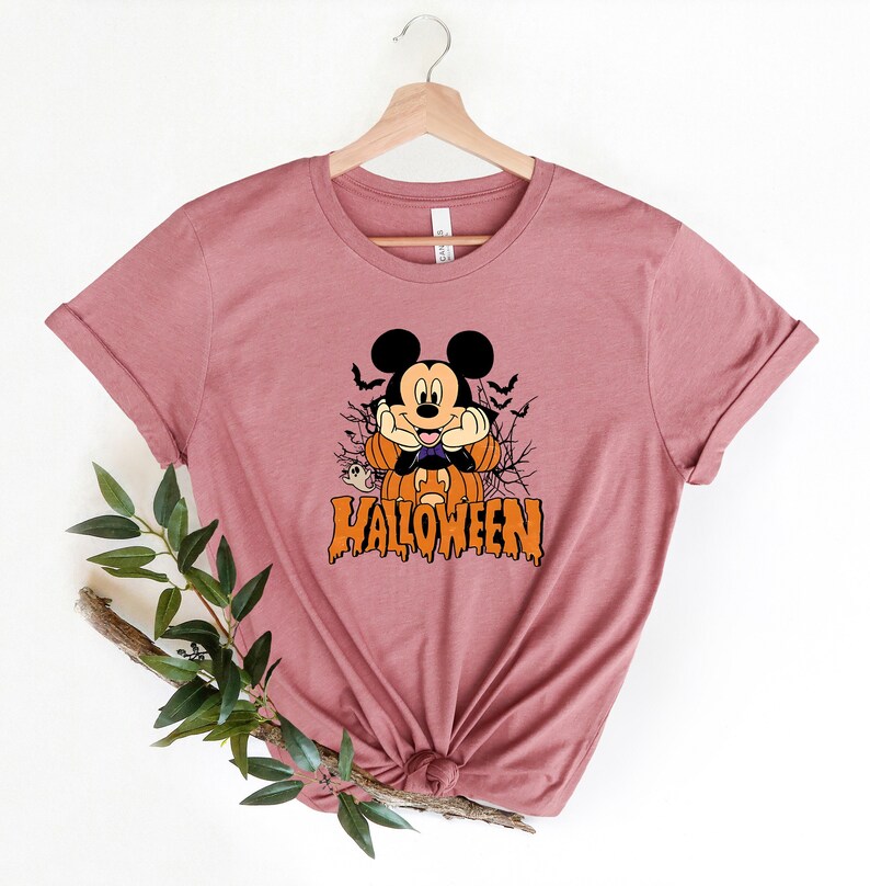 K&ouml;nnte beinhalten: Ein mauvefarbenes T-Shirt mit einem Aufdruck von Mickey Mouse, der als Vampir verkleidet ist und einen K&uuml;rbis h&auml;lt. Der Text "HALLOWEEN" ist unter Mickey Mouse aufgedruckt.