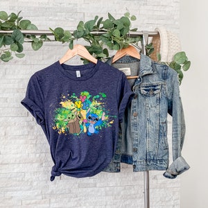 K&ouml;nnte beinhalten: Ein marineblaues T-Shirt mit einem bunten Farbklecks-Design, das Stitch und Baby Yoda zeigt, die Ballons halten.