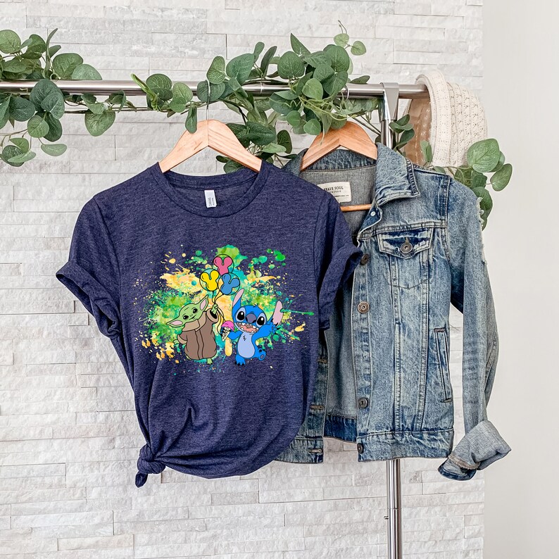 K&ouml;nnte beinhalten: Ein marineblaues T-Shirt mit einem bunten Farbklecks-Design, das Stitch und Baby Yoda zeigt, die Ballons halten.