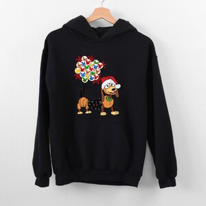 Pu&ograve; includere: Felpa con cappuccio nera con un cane dei cartoni animati che indossa un cappello da Babbo Natale e tiene delle luci natalizie. Il cane tiene anche un gruppo di ornamenti colorati. Un capo d'abbigliamento festivo a tema natalizio.