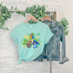 K&ouml;nnte beinhalten: Ein hellblaues T-Shirt mit einem Grafikdruck von Stitch und Baby Yoda, die Ballons halten. Die Grafik ist von gelben und gr&uuml;nen Farbspritzern umgeben.