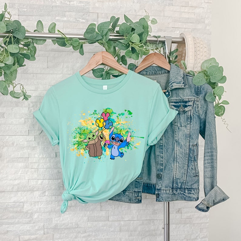 K&ouml;nnte beinhalten: Ein hellblaues T-Shirt mit einem Grafikdruck von Stitch und Baby Yoda, die Ballons halten. Die Grafik ist von gelben und gr&uuml;nen Farbspritzern umgeben.