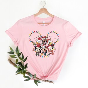May include: Pink t-shirt with a Christmas-themed design featuring Mickey Mouse and friends. The design shows Mickey Mouse, Minnie Mouse, Donald Duck, Goofy, Pluto, and Daisy Duck wearing Santa hats and surrounded by Christmas lights in the shape of Mickey Mouse's ears.