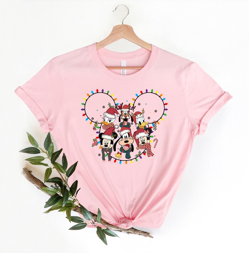 May include: Pink t-shirt with a Christmas-themed design featuring Mickey Mouse and friends. The design shows Mickey Mouse, Minnie Mouse, Donald Duck, Goofy, Pluto, and Daisy Duck wearing Santa hats and surrounded by Christmas lights in the shape of Mickey Mouse's ears.