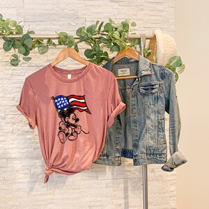 Puede incluir: Una camiseta rosa con una imagen de dibujos animados de Mickey Mouse sosteniendo una bandera estadounidense. La camiseta est&aacute; colgada en una percha con una chaqueta vaquera.